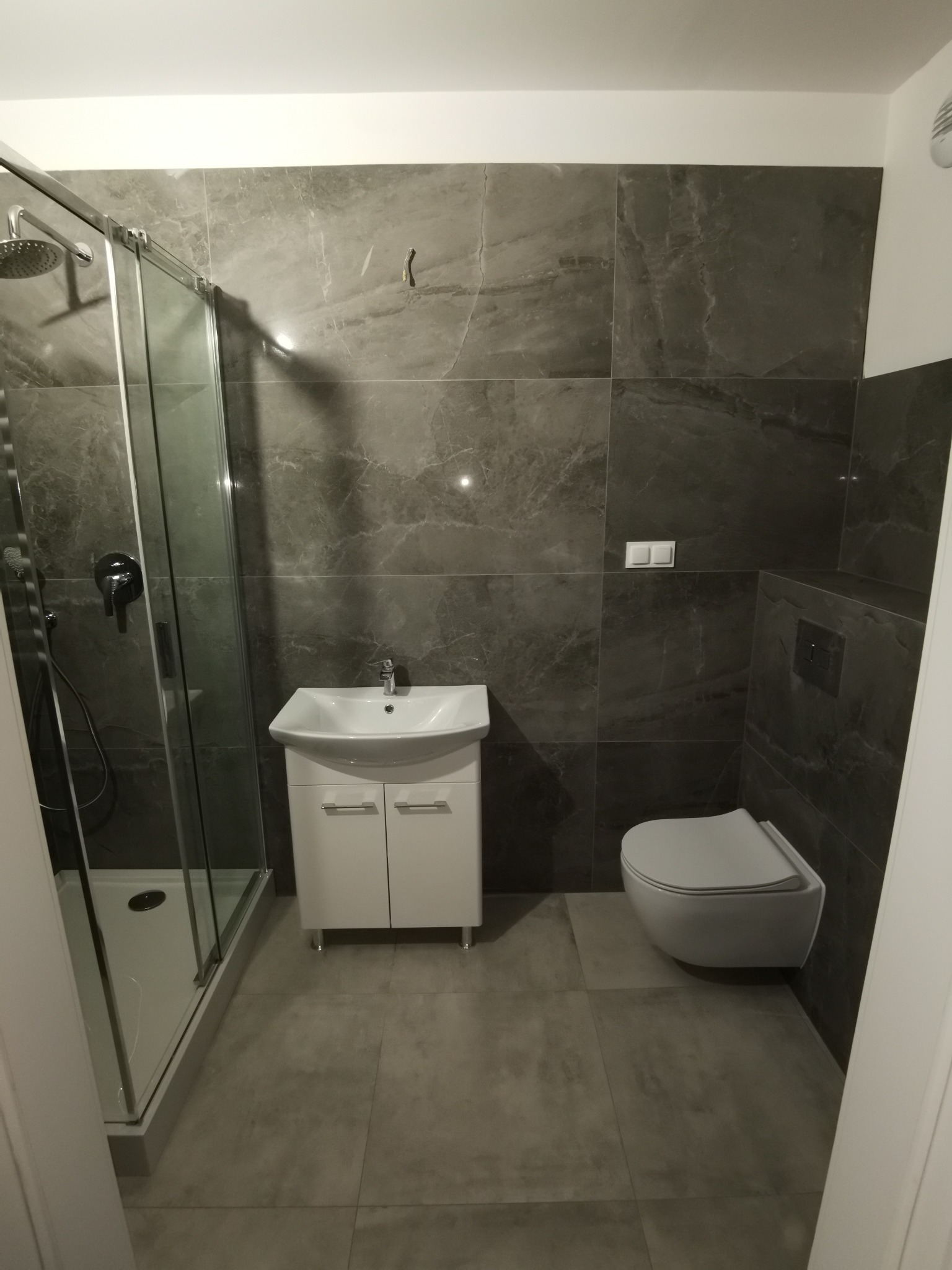 Nowoczesna łazienka z szarymi płytkami imitującymi marmur, kabina prysznicowa ze szklanymi drzwiami przesuwnymi, podwieszana umywalka z szafką i wisząca toaleta.