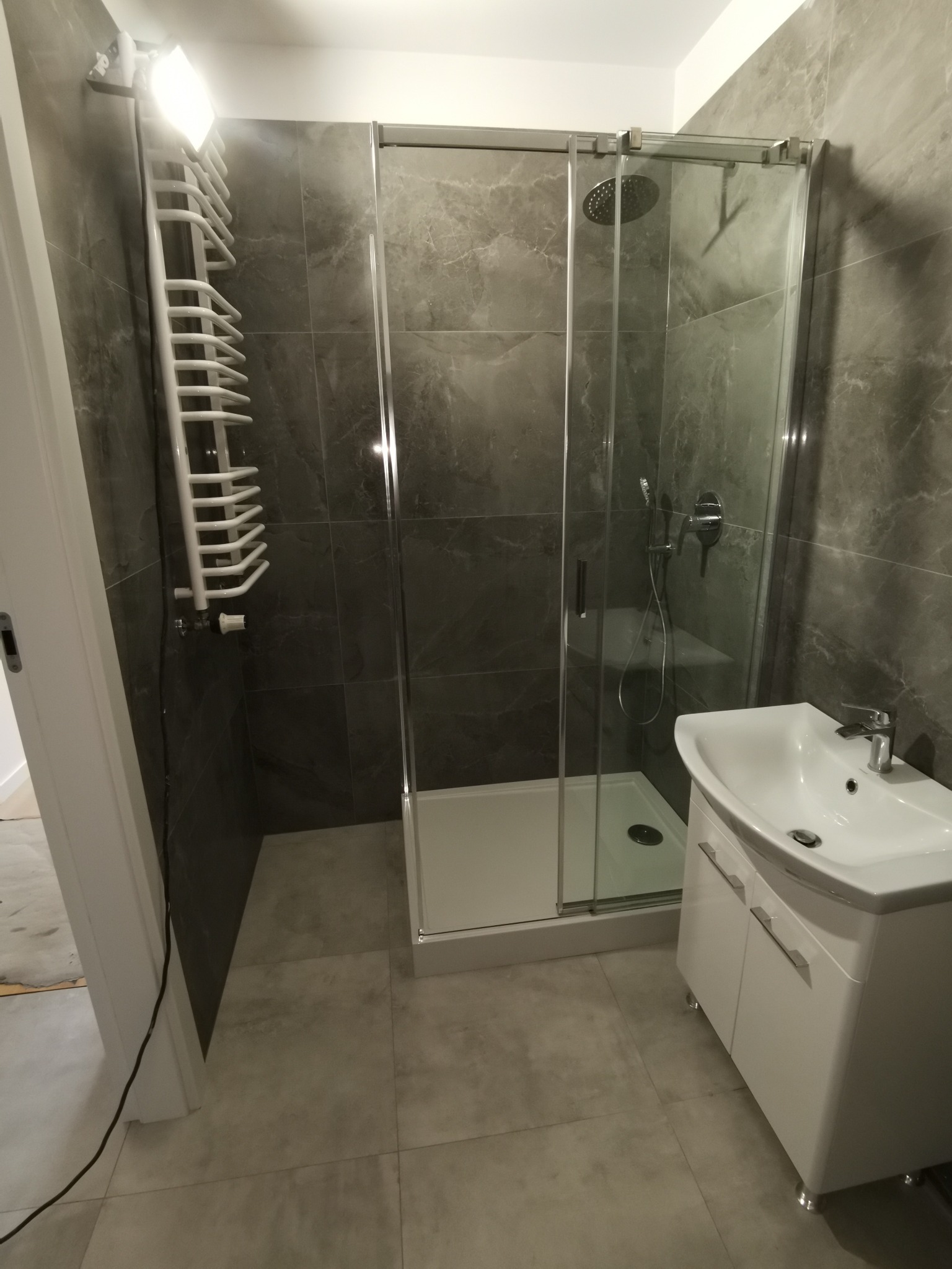 Nowoczesna łazienka z szarymi płytkami imitującymi marmur, kabina prysznicowa z przezroczystego szkła, biała umywalka z szafką i grzejnik drabinkowy.