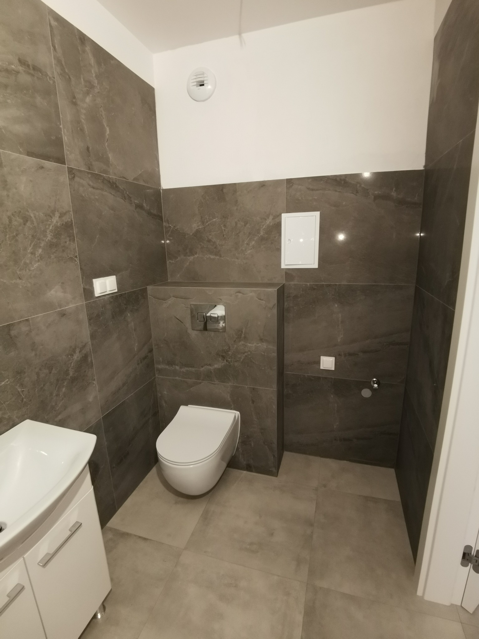 Nowoczesna łazienka wykończona w szarościach, z wiszącą toaletą, umywalką z szafką i dużymi płytkami imitującymi kamień na ścianach i podłodze. Widoczna biała wentylacja sufitowa i klapka rewizyjna.