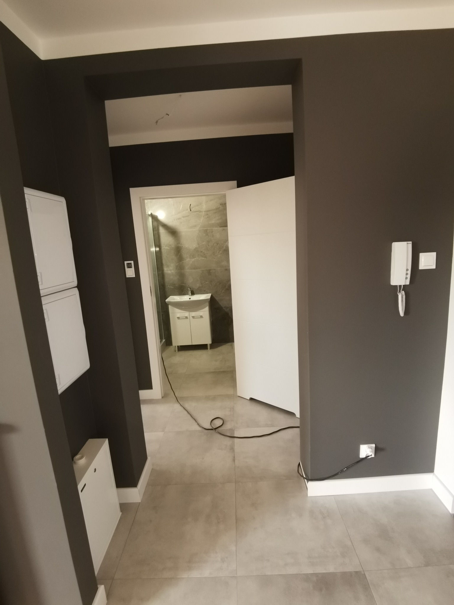 Widok na łazienkę z umywalką i szarą glazurą przez otwarte białe drzwi, ściany w korytarzu pomalowane na ciemny szary kolor, widoczny biały domofon i panele elektryczne po lewej stronie.