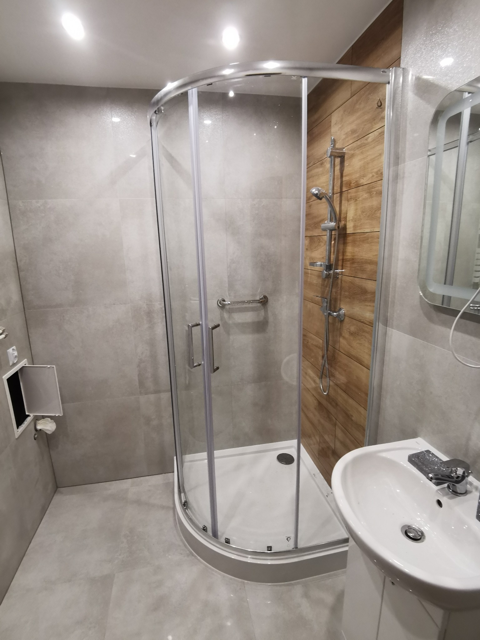 Nowoczesna łazienka z kabiną prysznicową z zaokrąglonymi szybami, płytkami imitującymi drewno pod prysznicem i umywalką na postumencie, wykończona w odcieniach szarości.