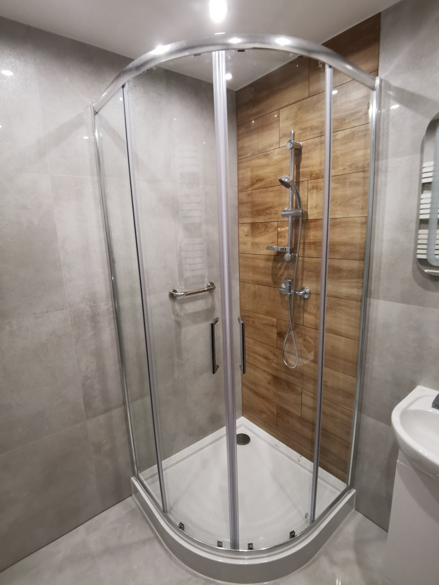 Kabina prysznicowa z chromowanymi profilami i uchwytem, drewnopodobne płytki na ścianie wewnątrz, szare płytki na ścianach i podłodze, biała umywalka w rogu.