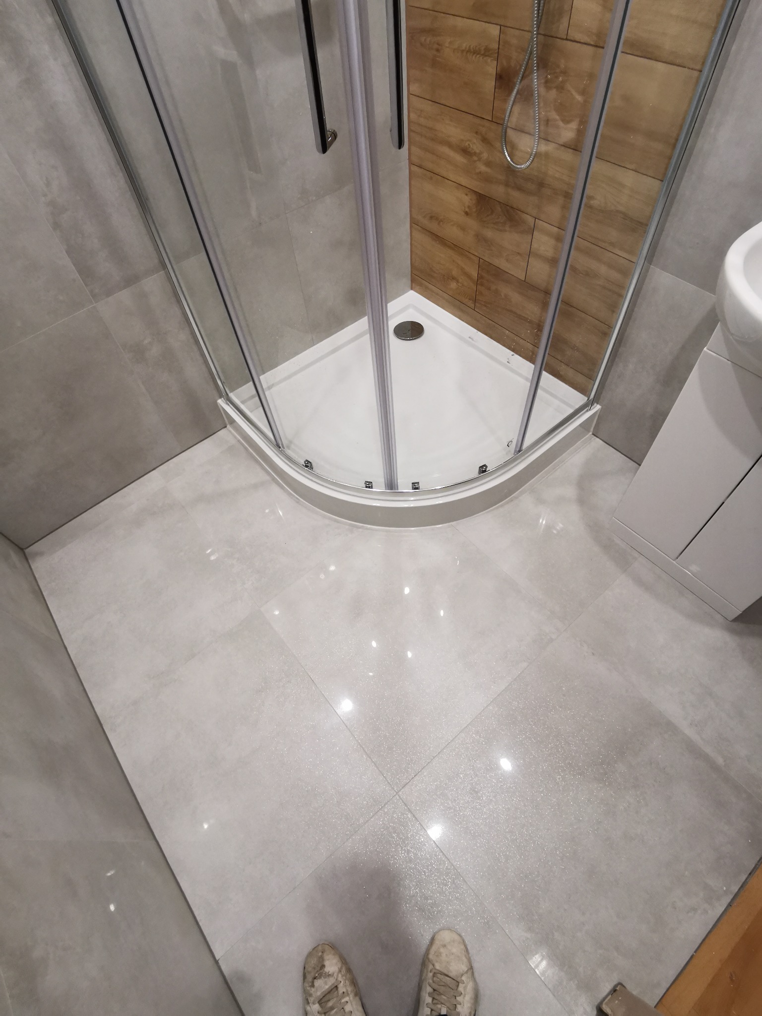 Nowoczesna łazienka po remoncie z narożną kabiną prysznicową z chromowanymi profilami, drewnopodobnymi płytkami wewnątrz kabiny, szarą płytką podłogową z połyskiem i butami w dolnym kadrze.