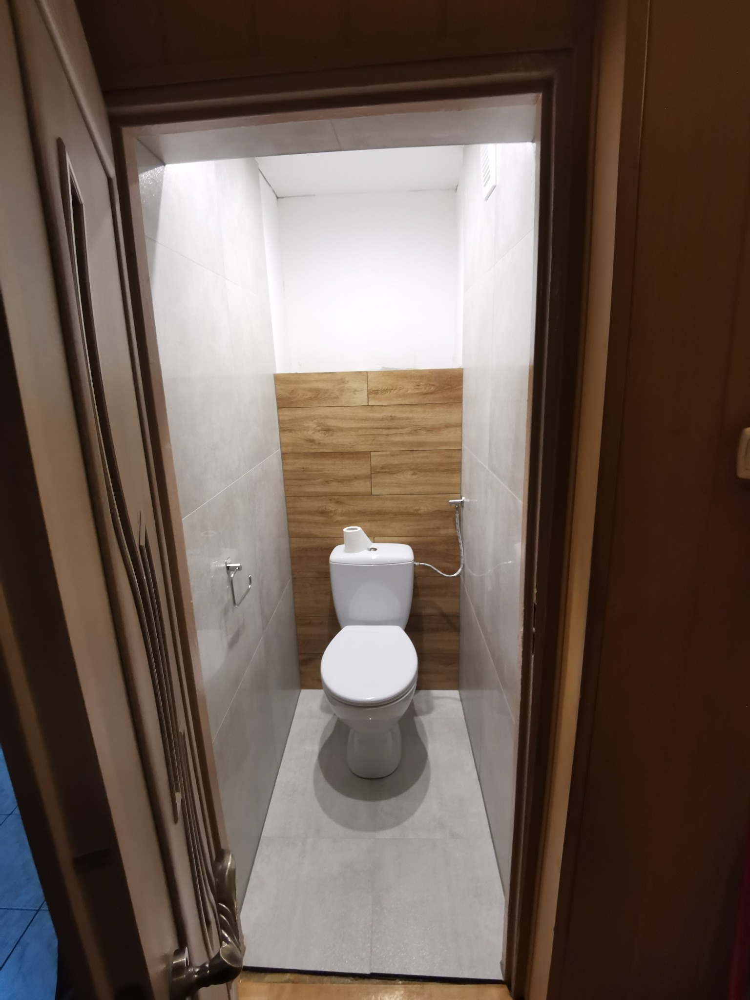 Wnętrze małej toalety po remoncie, widok zza uchylonych, zdobionych drzwi. Ściany wyłożone szarymi płytkami, fragment ściany nad toaletą w okładzinie imitującej drewno, biała toaleta kompaktowa...