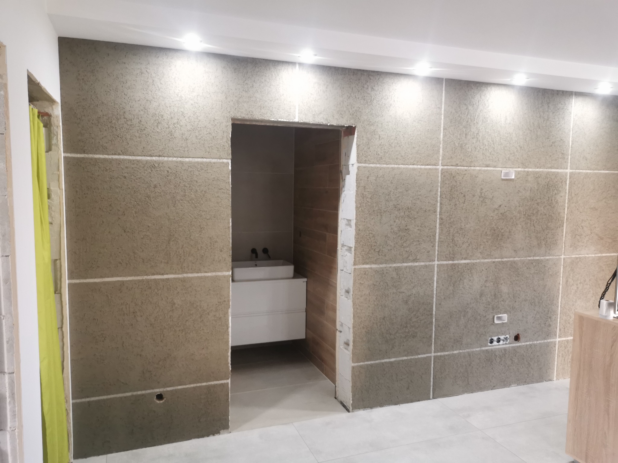 Ściana wykończona tynkiem strukturalnym w neutralnym odcieniu, podzielona na kwadratowe panele białymi listwami. Widoczne gniazdka elektryczne i otwór na instalację. W tle fragment łazienki z białą...