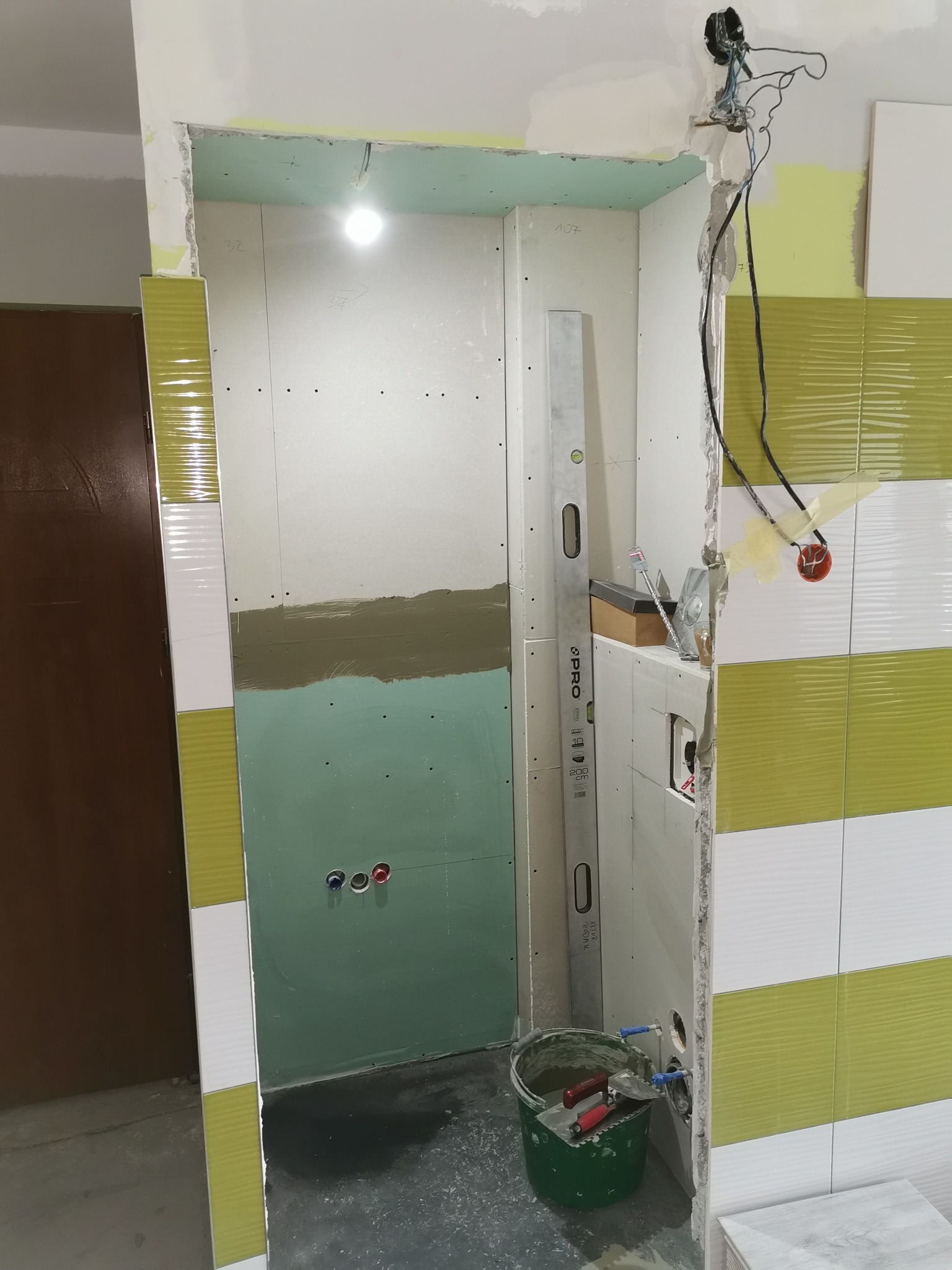 Wnętrze łazienki w trakcie prac wykończeniowych, widoczne płyty gipsowo-kartonowe, fragment ułożonych płytek w odcieniach bieli i limonki, poziomica oparta o ścianę, wiadro z narzędziami...
