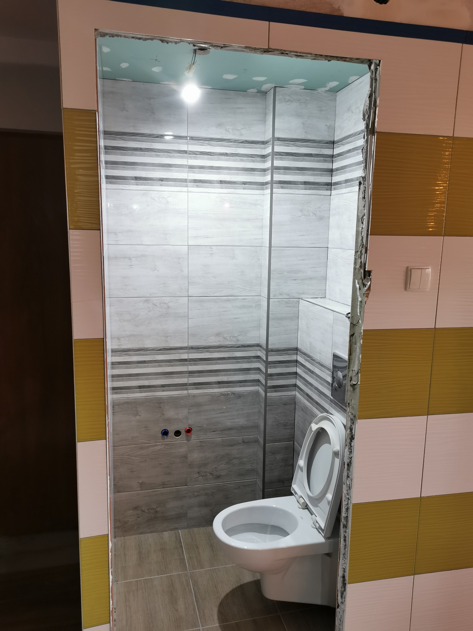 Wnętrze łazienki w trakcie remontu z widoczną toaletą, surowymi krawędziami drzwi i płytkami w odcieniach szarości i bieli oraz żółci, z widocznymi przyłączami do baterii.