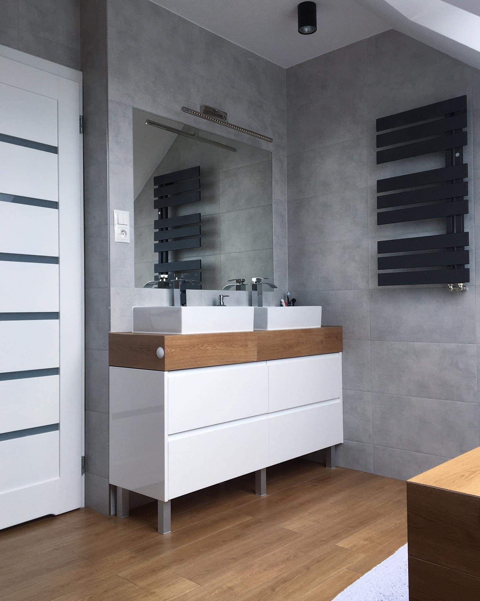 Nowoczesna łazienka z szarymi płytkami, białymi szafkami pod umywalki z drewnianym blatem, czarnym grzejnikiem drabinkowym i białymi drzwiami z przeszkleniami.