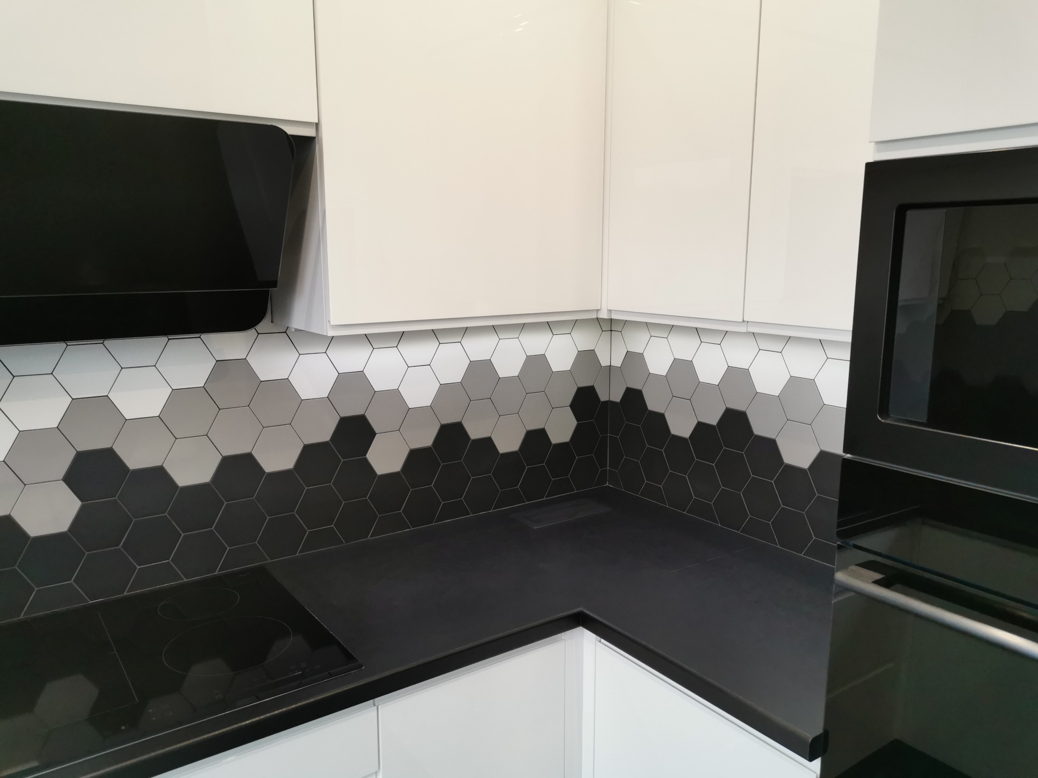Fragment nowoczesnej kuchni z białymi szafkami, czarnym blatem i heksagonalnymi płytkami w odcieniach szarości na ścianie, widoczny czarny okap i piekarnik.