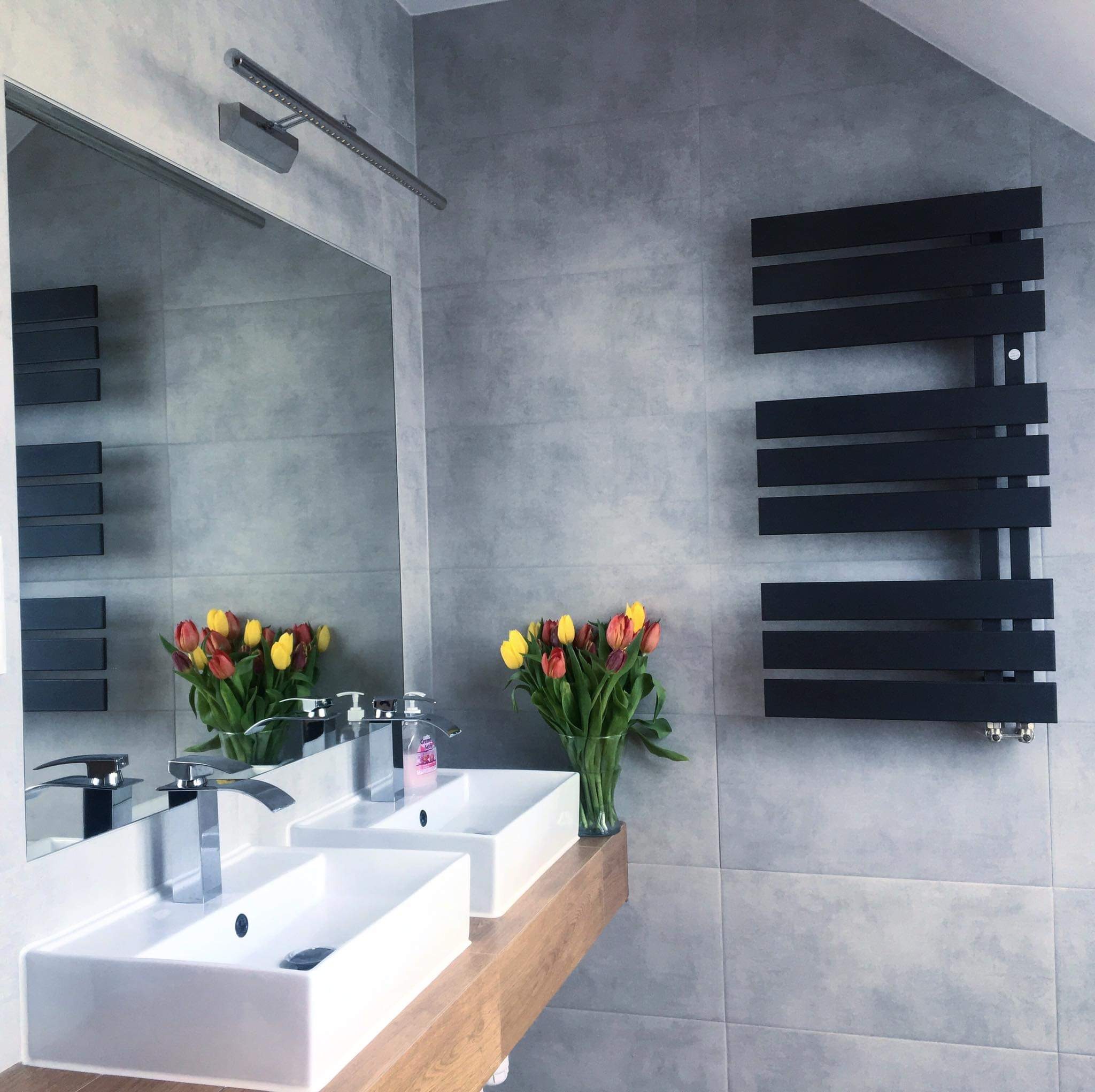 Nowoczesna łazienka z dwiema białymi umywalkami nablatowymi na drewnianym blacie, dużym lustrem z oświetleniem LED, czarnym grzejnikiem drabinkowym na szarej ścianie z płytek oraz bukietem tulipanów.