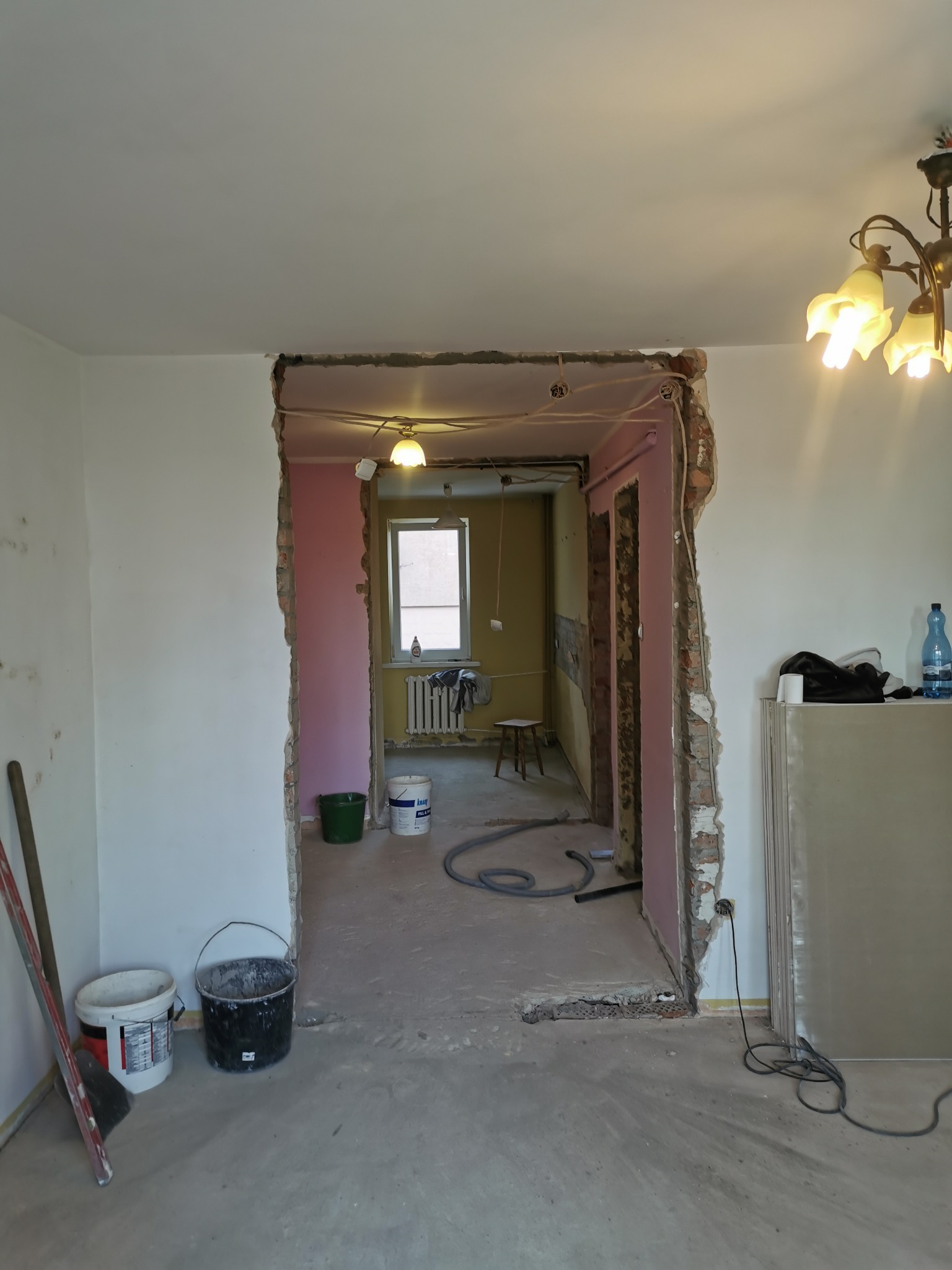 Przygotowania do prac wykończeniowych: widoczne ślady po kuciu ścian, odkryte przewody elektryczne, wiadro z zaprawą i płyty gipsowo-kartonowe w pomieszczeniu przed remontem.