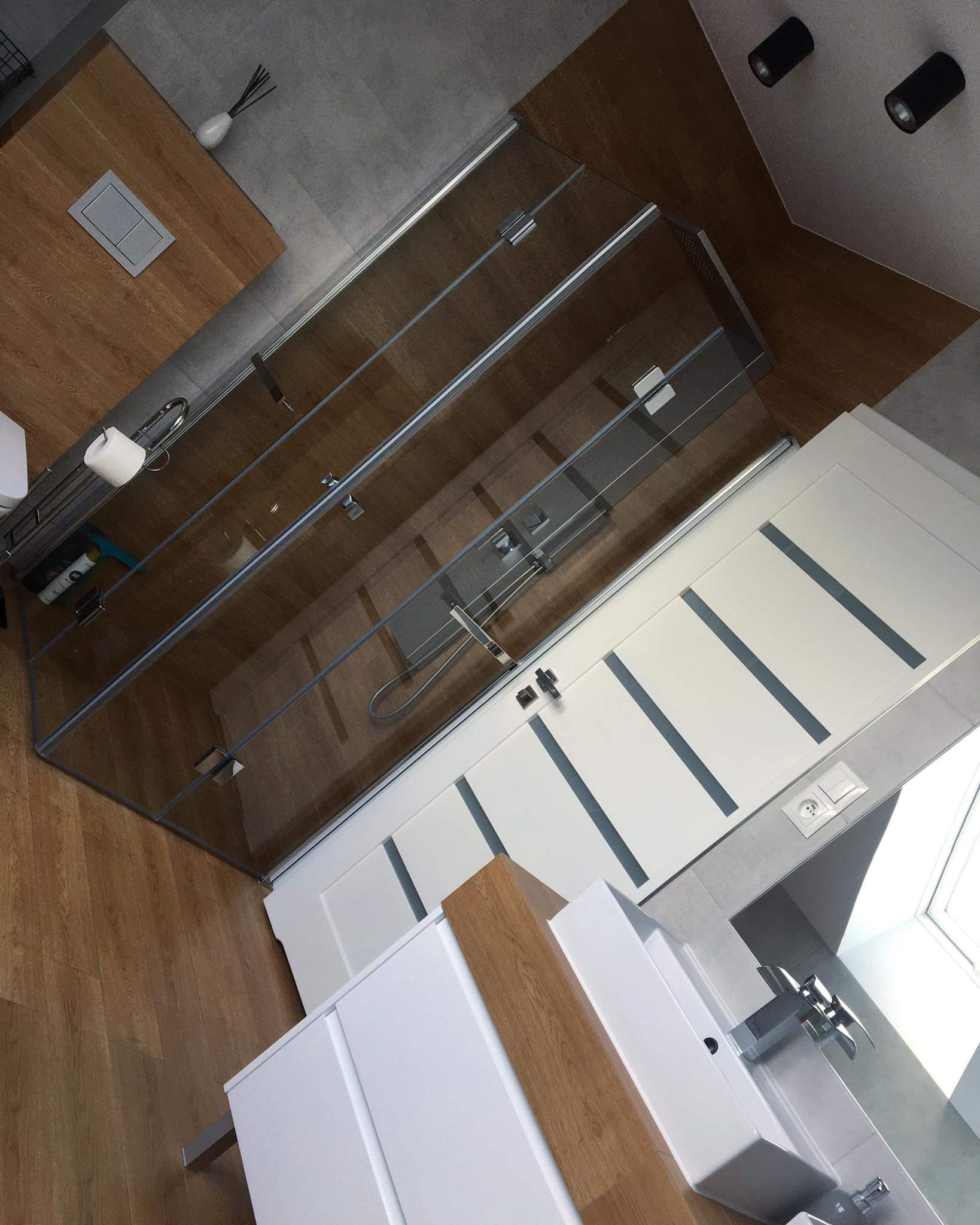 Nowoczesna łazienka z kabiną prysznicową ze szklanymi drzwiami, białą szafką z umywalką i drewnianymi akcentami, oświetlona punktowymi lampami sufitowymi.