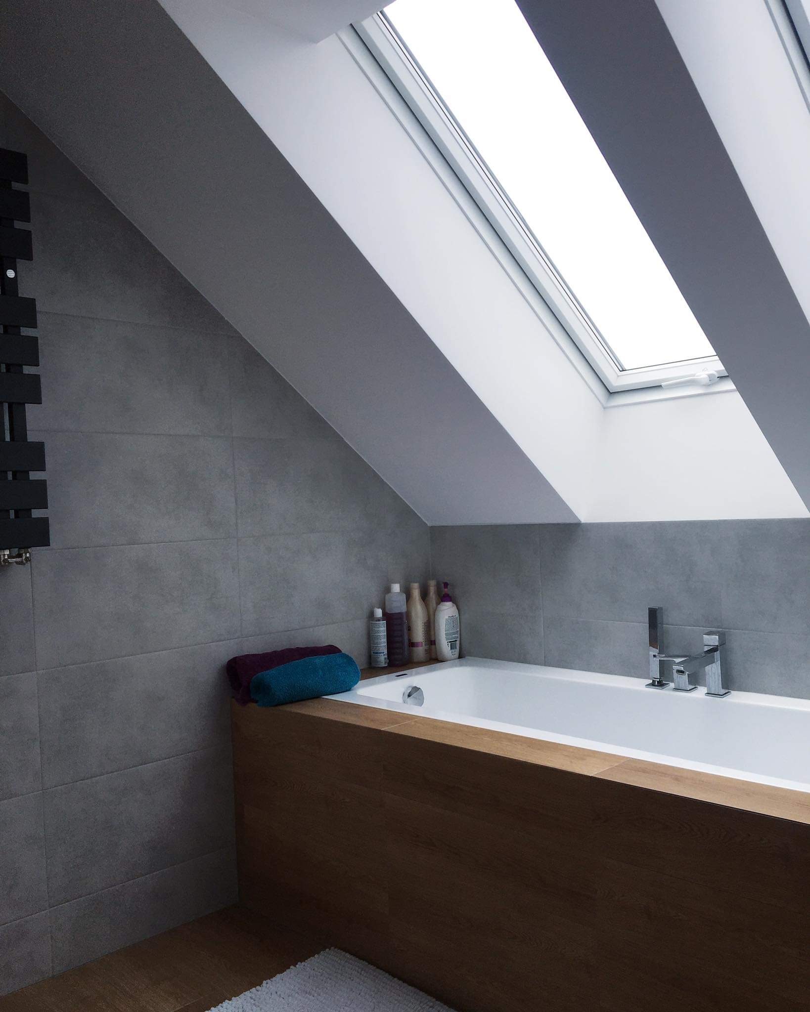 Nowoczesna łazienka na poddaszu z wanną obudowaną drewnem, szarymi płytkami i oknem dachowym, doświetlającym wnętrze.