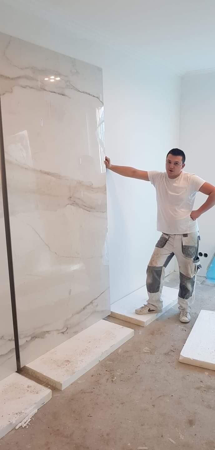 Instalator opiera się o dużą, błyszczącą płytę imitującą marmur, ustawioną pionowo na podłodze wyłożonej styropianem w trakcie prac wykończeniowych w jasnym pomieszczeniu.