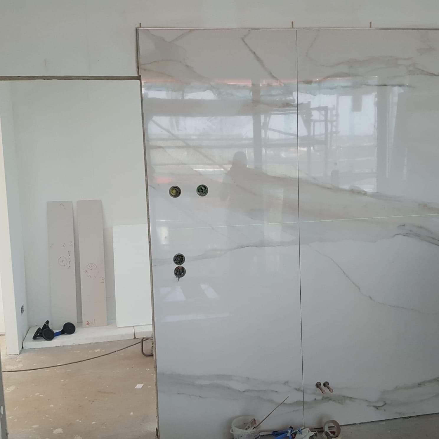 Płytki imitujące marmur na ścianie w trakcie montażu w łazience, widoczne otwory pod gniazdka elektryczne, na podłodze narzędzia i resztki materiałów budowlanych, w tle płyty kartonowo-gipsowe.