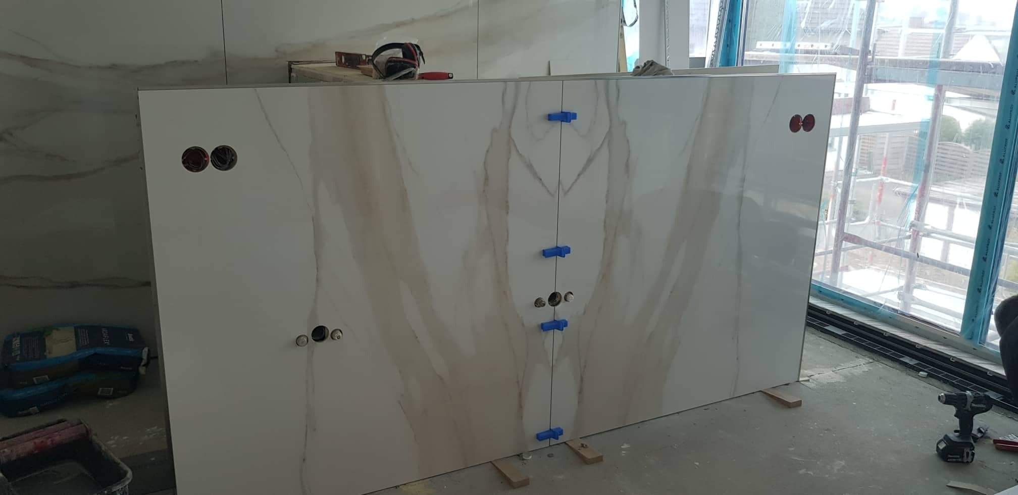 Montaż dużych, marmurkowych płytek ściennych w łazience z użyciem niebieskich krzyżyków dystansowych i poziomicy, widoczne otwory na gniazdka elektryczne.