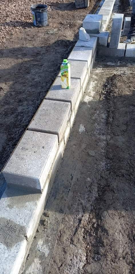 Wznoszenie fundamentu z betonowych bloczków, na wierzchu karton soku, widoczny mokry grunt i wiadro w tle.