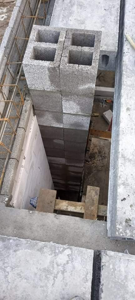 Wznoszenie pionowego komina z szarych bloczków betonowych z widocznym zbrojeniem wewnątrz i drewnianym szalunkiem u podstawy, widok z góry na placu budowy.