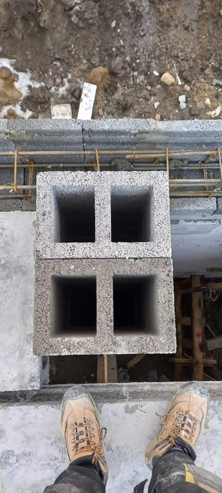 Dwa bloczki fundamentowe z czterema otworami ułożone na fundamencie z wystającym zbrojeniem, widok z góry na buty robotnika.