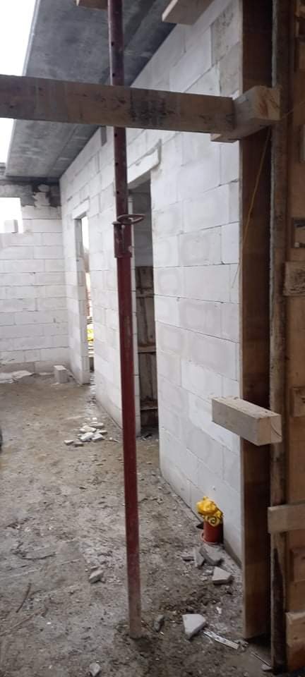 Wnętrze domu w trakcie budowy z białych bloczków, widoczne podpory stropu, drewniane szalunki ościeżnicy i resztki materiałów budowlanych na podłożu.