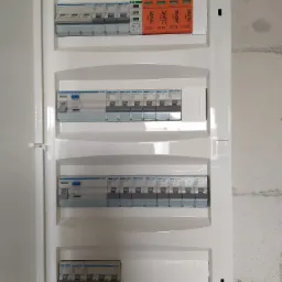 Rozdzielnica elektryczna z zamontowanymi bezpiecznikami i ogranicznikami przepięć marek Hager i Simtec w białej obudowie, widok z bliska na tle surowej ściany.