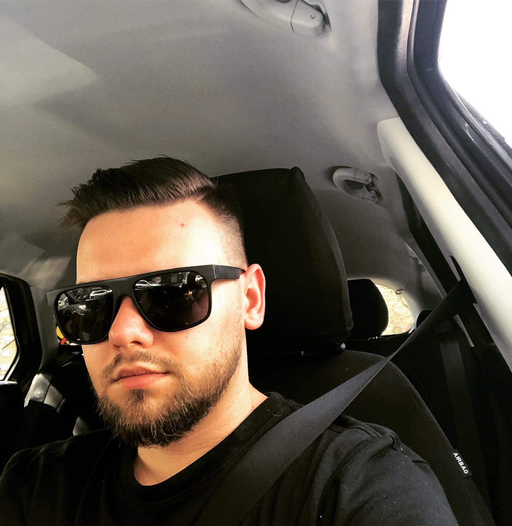 Mężczyzna w czarnych okularach siedzi w samochodzie, zapięty pasem bezpieczeństwa, widoczna deska rozdzielcza i wnętrze pojazdu.