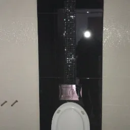 Nowoczesna toaleta z czarno-białymi płytkami; pionowy pas z mozaiki z połyskujących płytek na czarnym tle; widoczne mocowania na ścianie po lewej stronie.