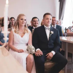 Młoda para siedzi obok siebie podczas ceremonii ślubnej, trzymając się za ręce, panna młoda w białej sukni trzyma bukiet, w tle widoczni goście.