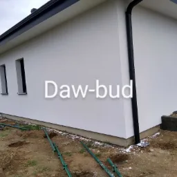 Elewacja domu jednorodzinnego po malowaniu na biało, widoczne czarne rynny i okna, teren wokół domu w trakcie prac ziemnych.