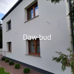 Dwukondygnacyjny dom z jasną elewacją i brązowymi oknami, widoczne ślady po naprawach na ścianie, ozdobne krzewy i kamyki wzdłuż fundamentów.