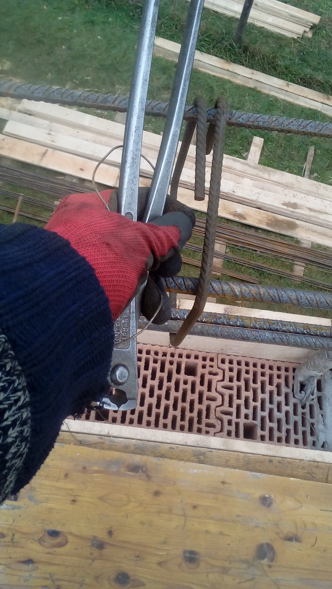 Ręka w czerwonej rękawicy wiąże drutem zbrojeniowym pręty zbrojeniowe za pomocą kombinerek Knipex, widoczny bloczek ceramiczny i deski szalunkowe.