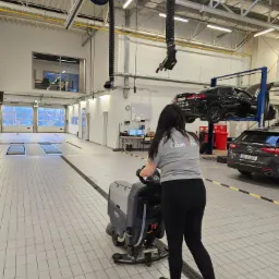 Kobieta w szarej koszulce z logo firmy sprzątającej obsługuje maszynę do czyszczenia podłóg w przestronnym warsztacie samochodowym z podnośnikami i samochodami. Jasne oświetlenie, białe ściany...