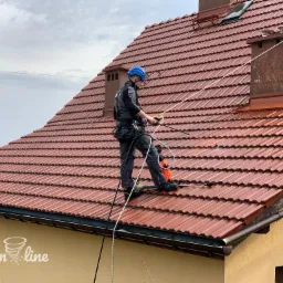 Pracownik w uprzęży, z niebieskim kaskiem, czyści czerwoną dachówkę za pomocą myjki ciśnieniowej. Część dachu jest już umyta, a na ścianie budynku widoczne logo firmy.