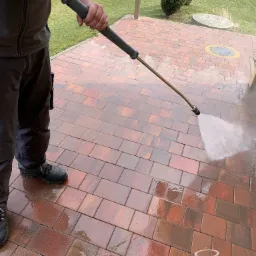 Mężczyzna czyści ciśnieniowo czerwoną kostkę brukową na podjeździe, widoczny spryskiwacz i woda pod ciśnieniem.