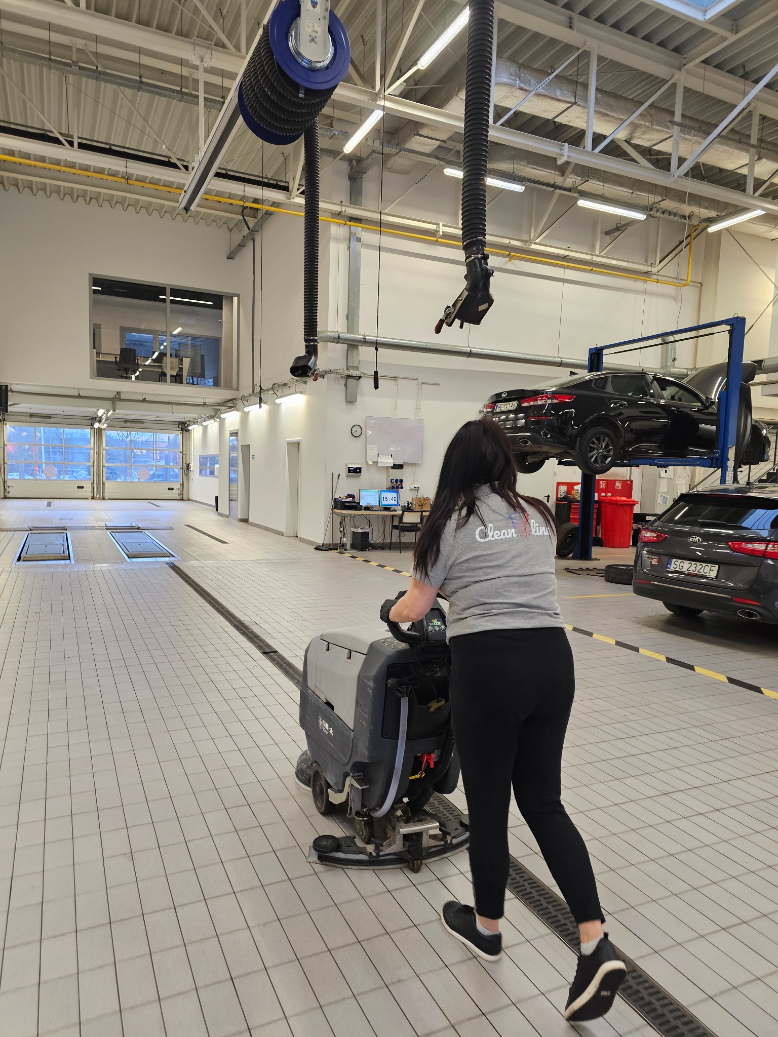 Kobieta w szarej koszulce z logo firmy sprzątającej obsługuje maszynę do czyszczenia podłóg w przestronnym warsztacie samochodowym z podnośnikami i samochodami. Jasne oświetlenie, białe ściany...