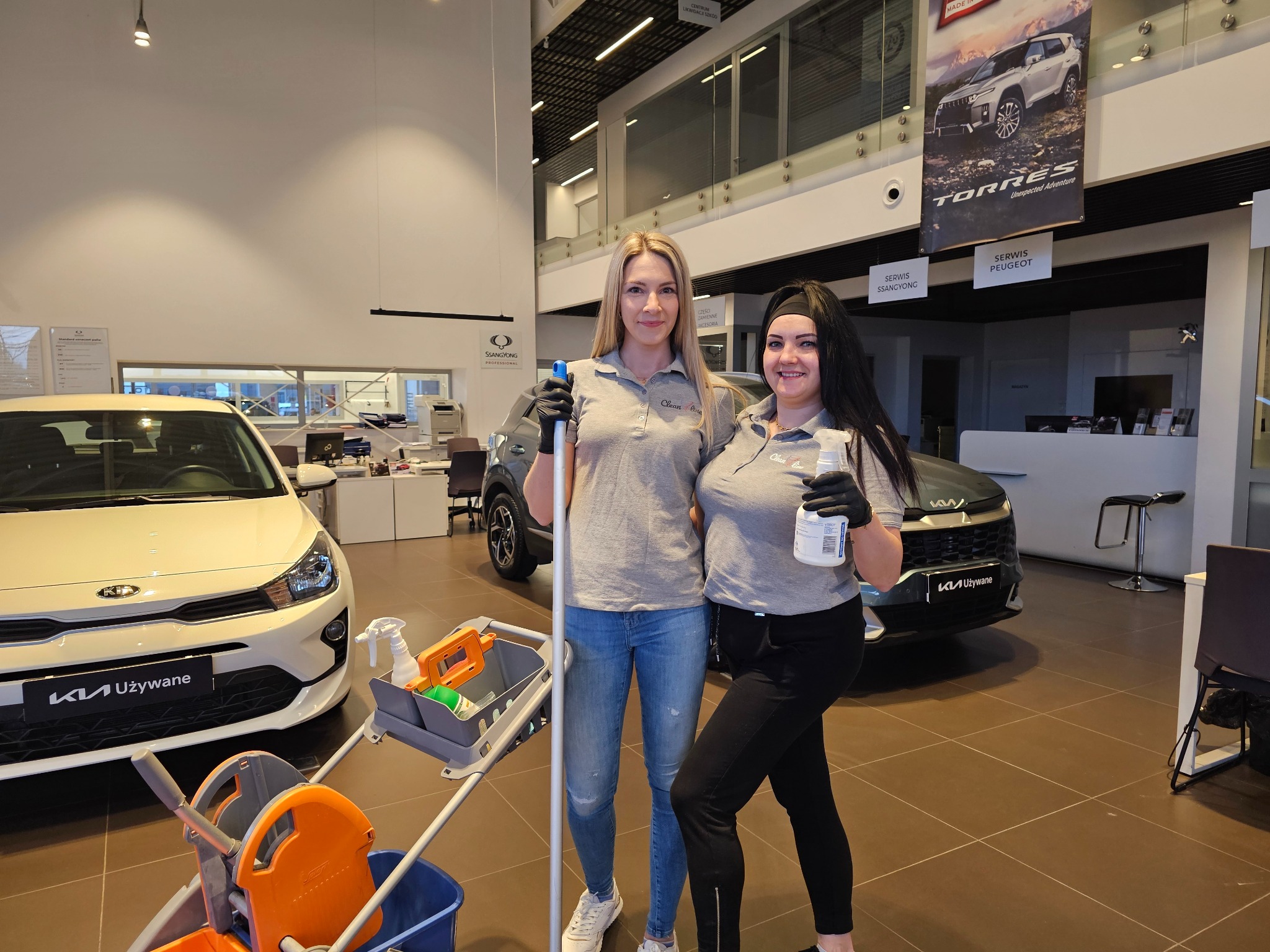 Dwie uśmiechnięte kobiety w szarych koszulkach z logo firmy sprzątającej, w czarnych rękawicach, stoją w salonie samochodowym z mopem i wózkiem do sprzątania, na tle samochodów marki Kia i plakatów...