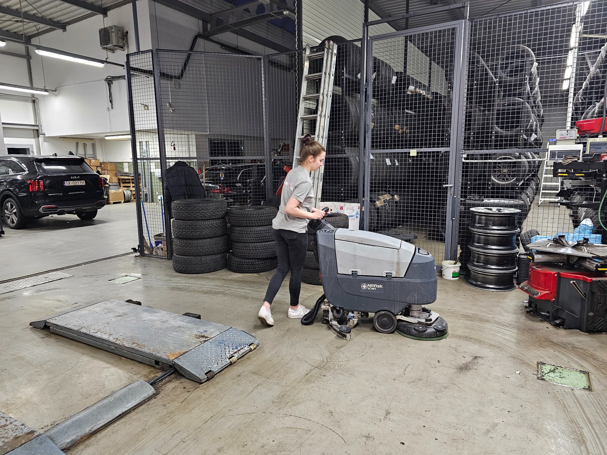 Kobieta obsługuje szorowarkę Nilfisk SC401 na betonowej posadzce warsztatu, w tle opony, felgi i podnośnik samochodowy.