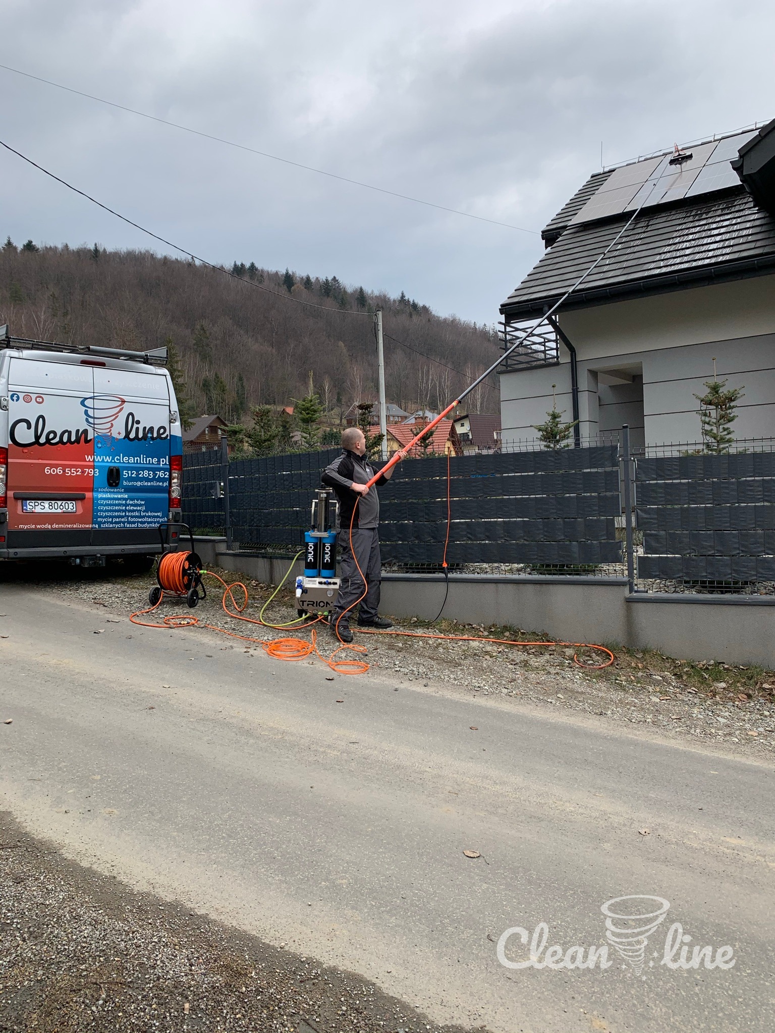 Pracownik firmy Clean Line czyści panele fotowoltaiczne na dachu domu jednorodzinnego za pomocą specjalistycznego wysięgnika i urządzenia ciśnieniowego. W tle widoczny bus firmy z danymi...