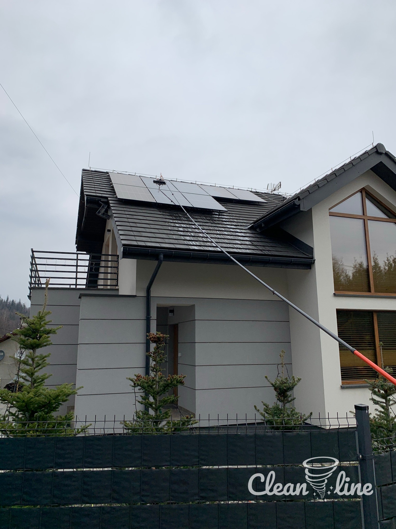 Czyszczenie paneli słonecznych na dachu domu jednorodzinnego za pomocą teleskopowej lancy z dyszą, widoczne logo firmy na ogrodzeniu.