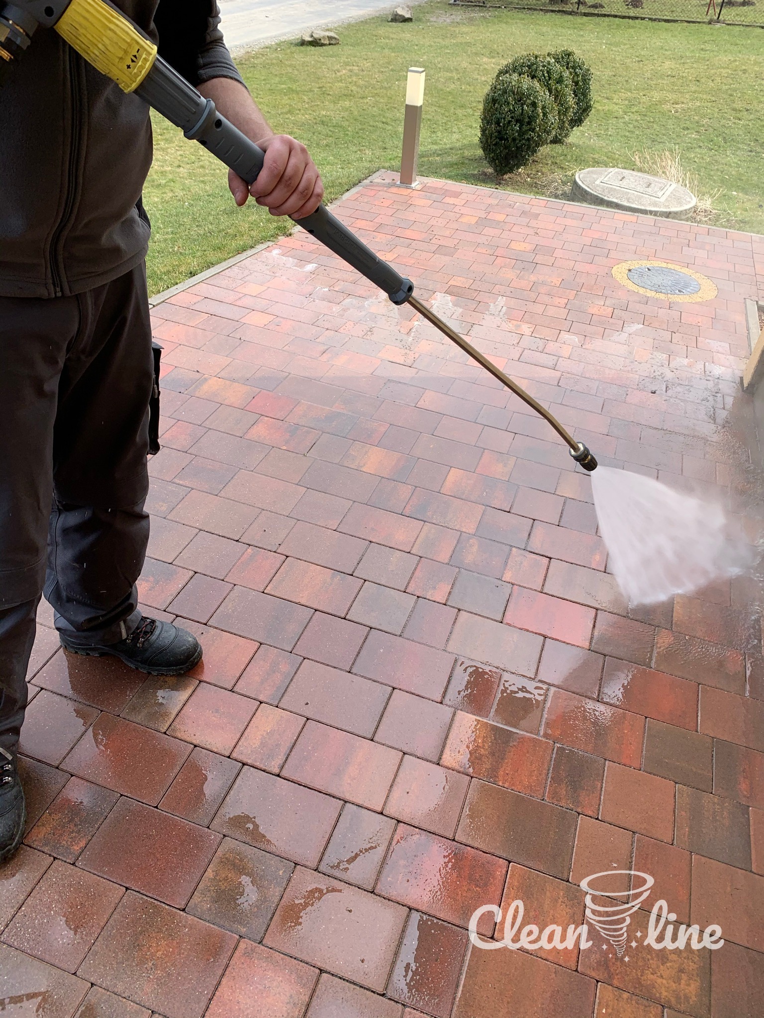 Mężczyzna czyści ciśnieniowo czerwoną kostkę brukową na podjeździe, widoczny spryskiwacz i woda pod ciśnieniem.
