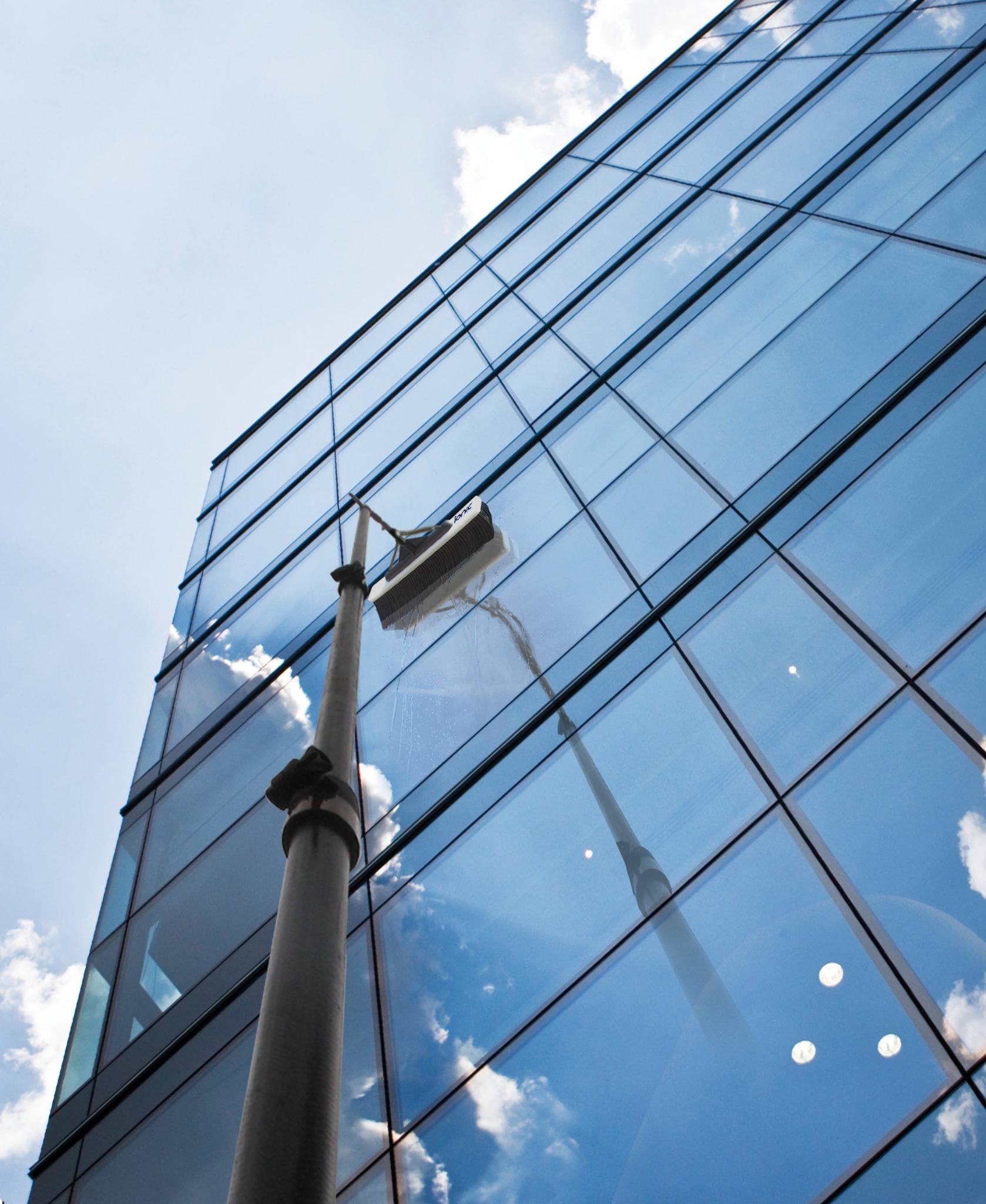 Mycie szklanej fasady budynku za pomocą szczotki teleskopowej; widoczne odbicie nieba i chmur na czyszczonej powierzchni.