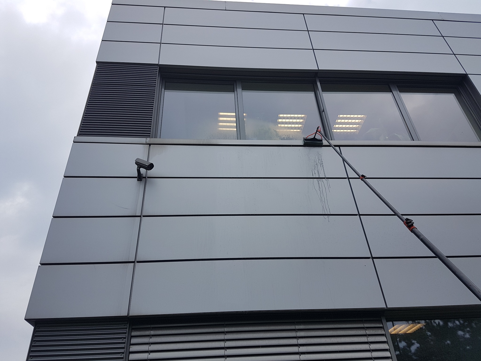 Czyszczenie okien na wysokości w budynku biurowym o szarej elewacji panelowej, widoczny wysięgnik z myjką i ślady spływającej wody.