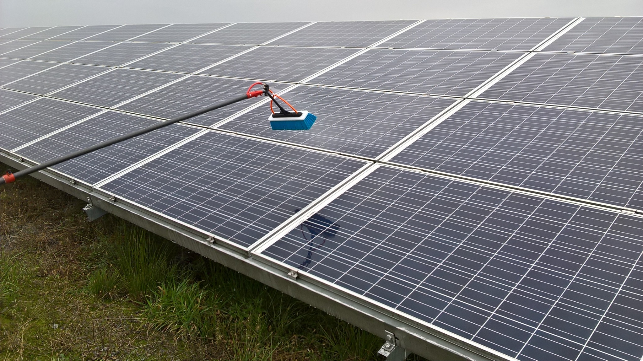 Czyszczenie paneli fotowoltaicznych za pomocą szczotki na długim wysięgniku, widoczne panele na metalowej konstrukcji i trawa w tle.