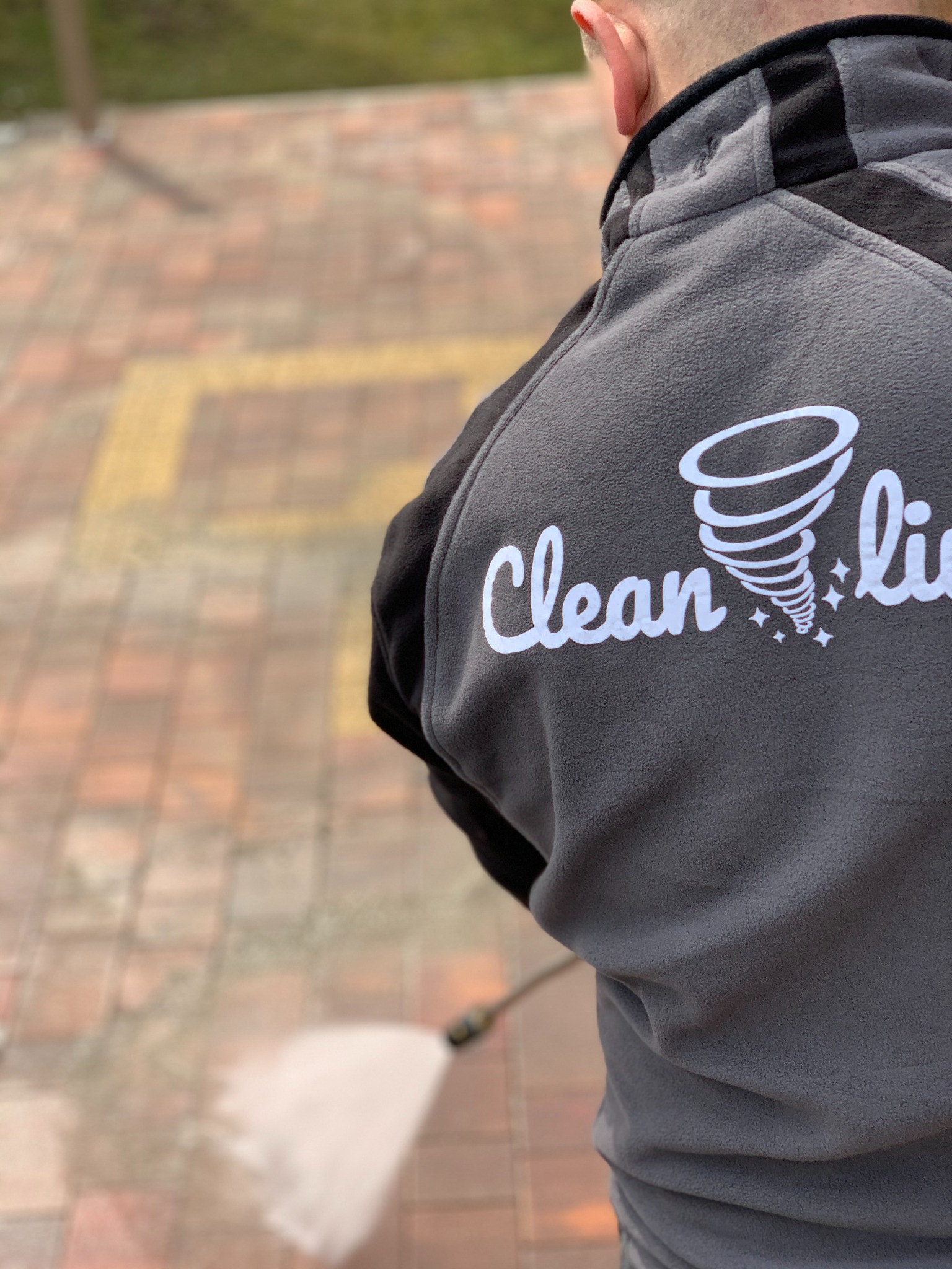 Pracownik w szarej bluzie z logo firmy 'Clean Line' czyści ciśnieniowo brukowaną nawierzchnię z zabrudzeń.
