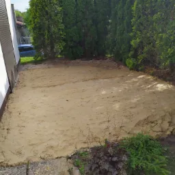 Przygotowany teren pod trawnik lub kostkę brukową, wyrównany piaskiem przy budynku i tujach.