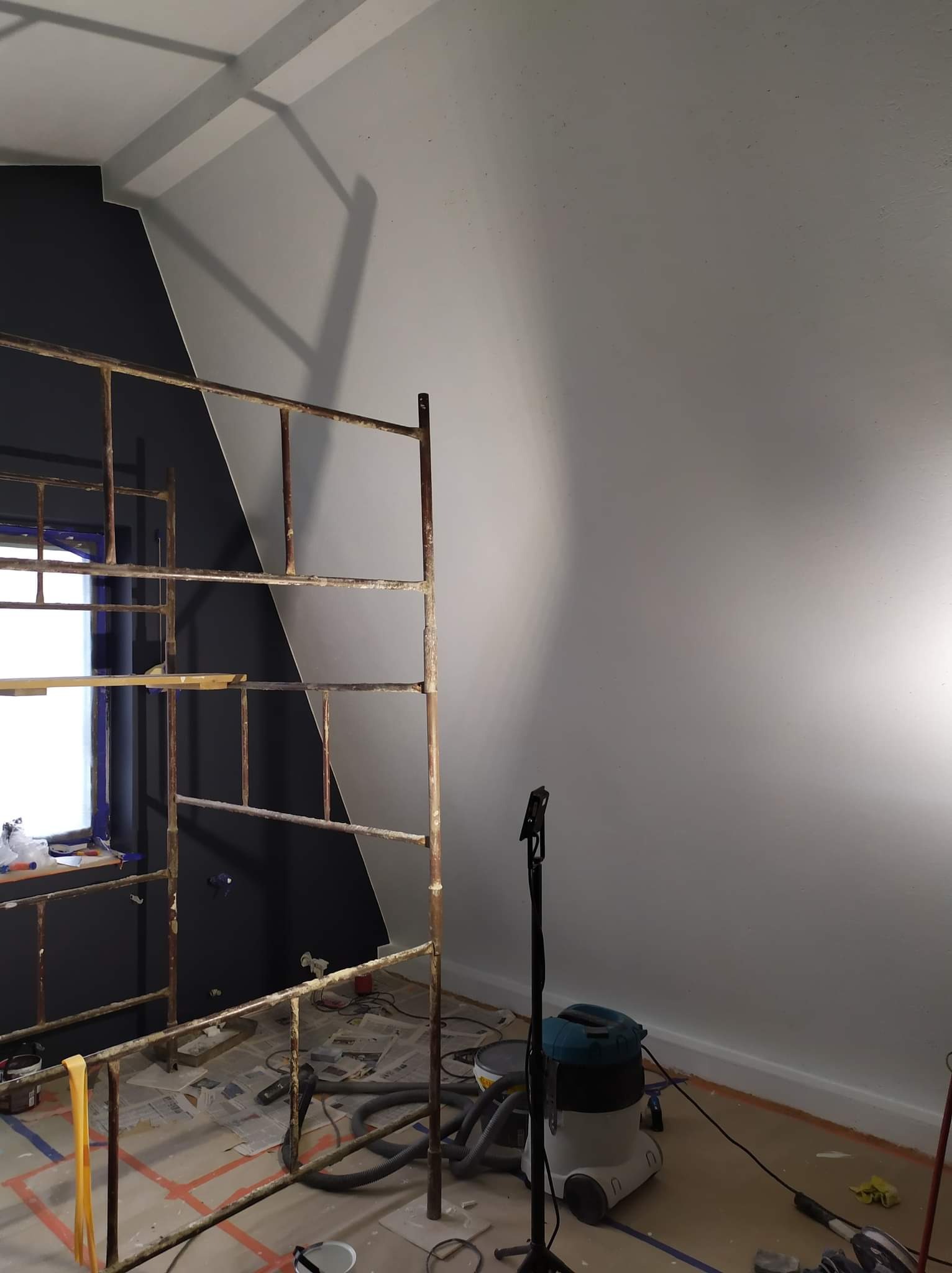Malowanie ścian w pokoju: ściana pomalowana częściowo na czarno i częściowo na szaro, widoczne rusztowanie, odkurzacz i zabezpieczenia podłogi.
