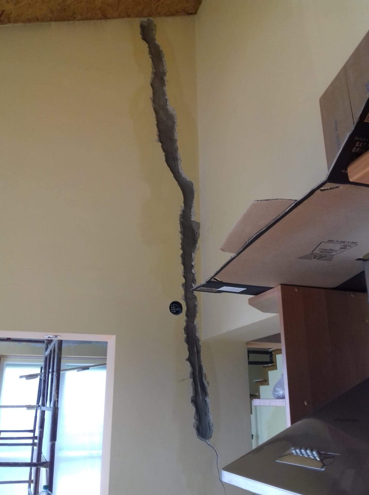 Pęknięcie pionowe na ścianie w trakcie naprawy, widoczne ślady po kuciu, fragment instalacji elektrycznej, w tle rusztowanie i fragment szafki kuchennej.