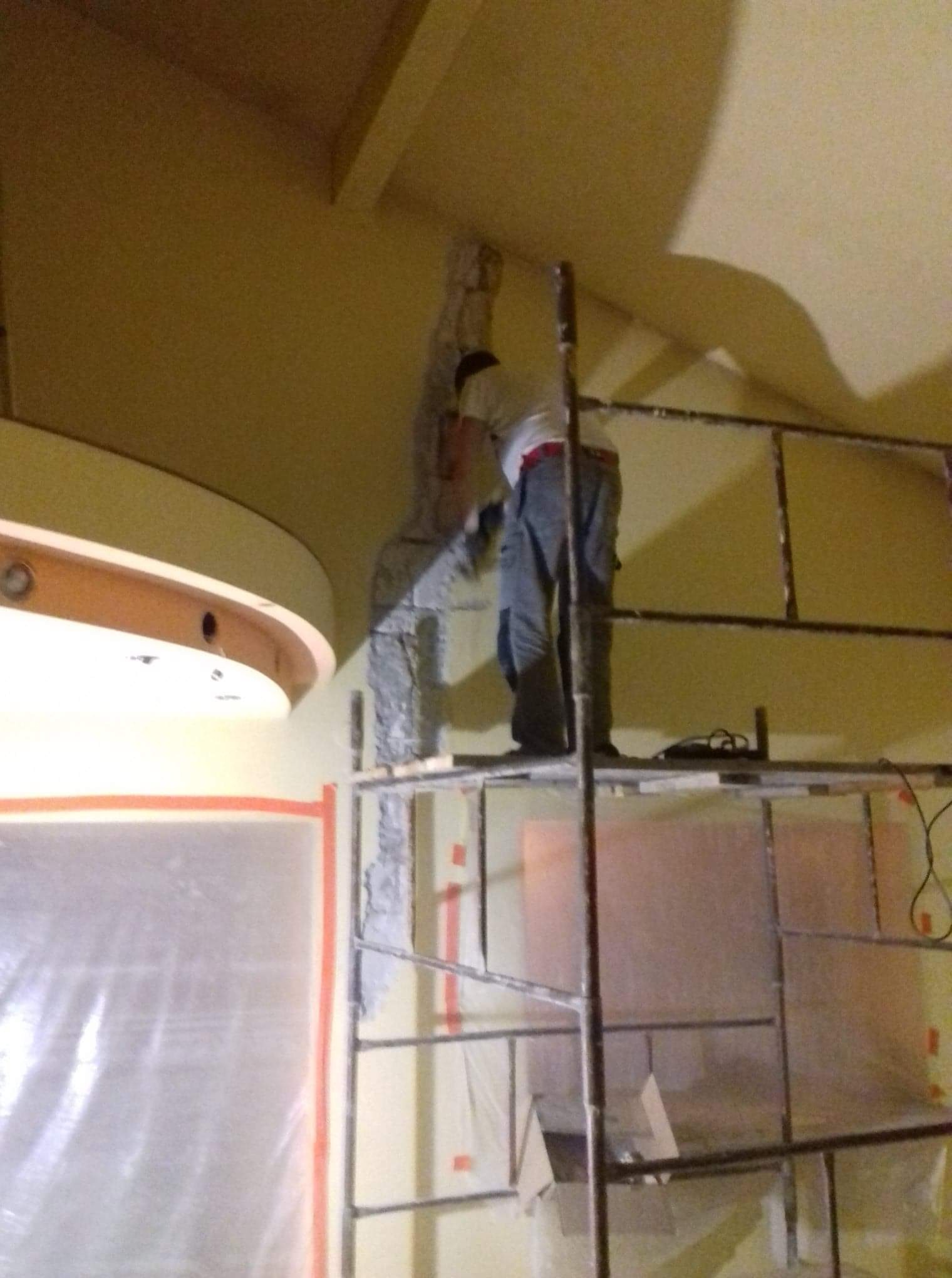 Pracownik na rusztowaniu skuwa tynk z filaru w pomieszczeniu przygotowanym do remontu, zabezpieczonym folią malarską.