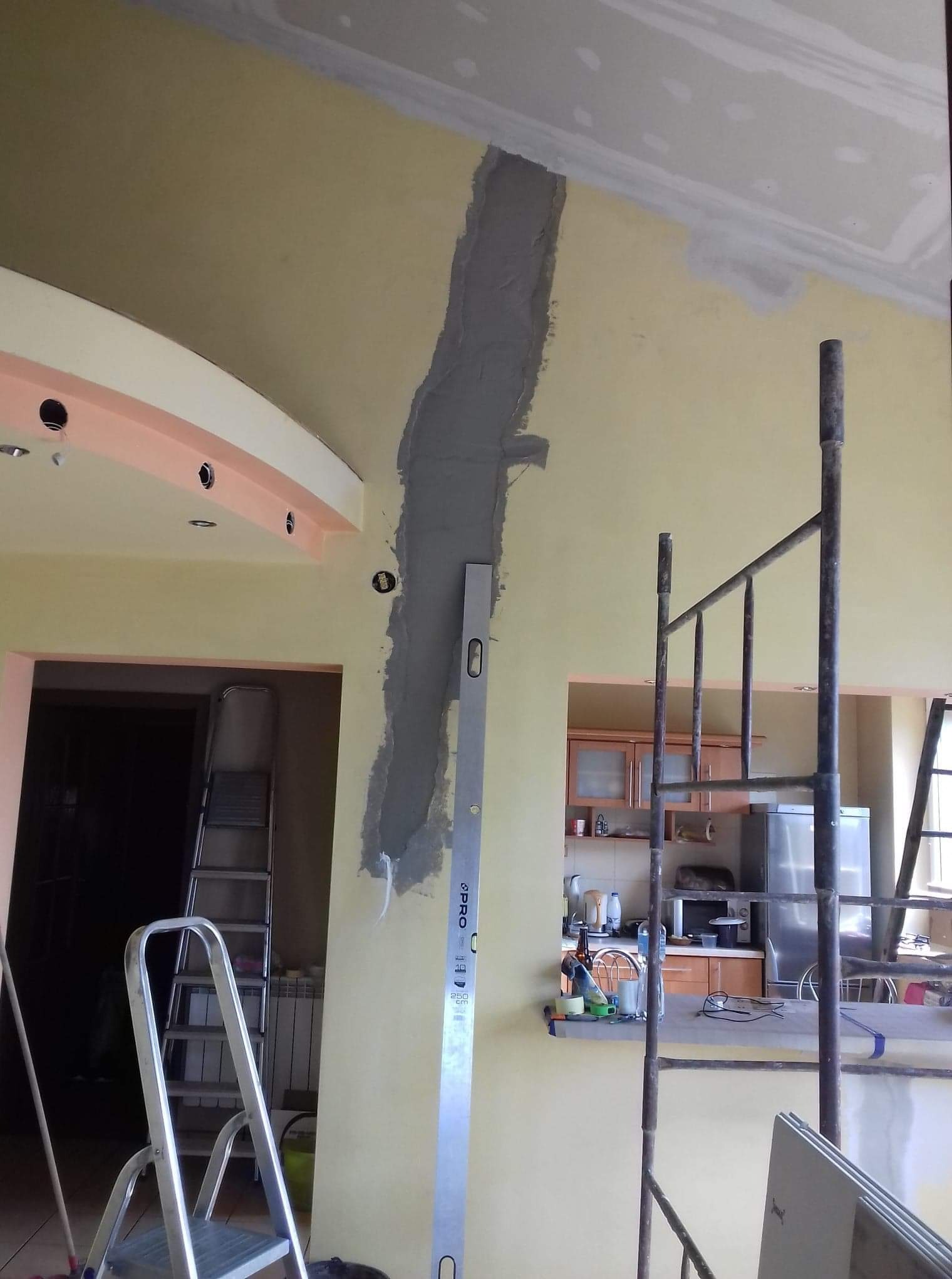 Wyrównywanie ściany gładzią w pionowym pasie, widoczna aluminiowa łata PRO i drabina, w tle otwarta kuchnia i rusztowanie