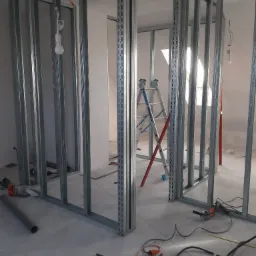 Wnętrze w trakcie budowy ścian działowych z metalowych profili, widoczna drabina, szlifierka kątowa i narzędzia na betonowej podłodze.